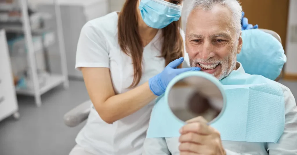 A masked dentist with gloves examines an elderly man’s teeth, who smiles while holding a mirror in a dental clinic.