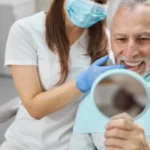 A masked dentist with gloves examines an elderly man’s teeth, who smiles while holding a mirror in a dental clinic. All-on-4 implant procedure