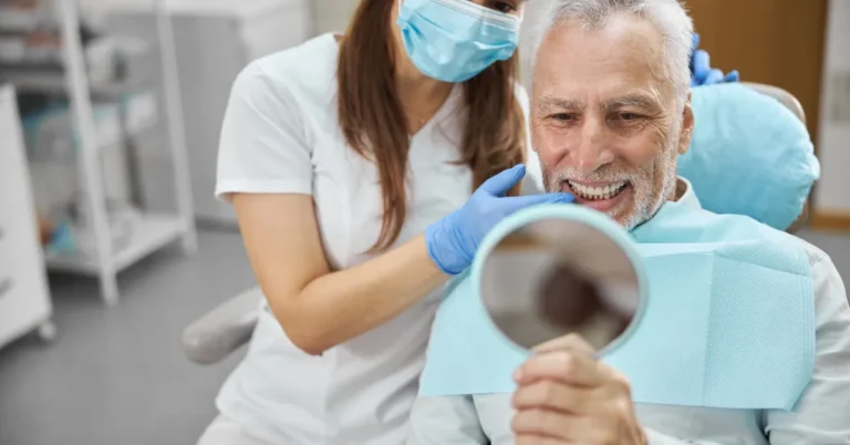 A masked dentist with gloves examines an elderly man’s teeth, who smiles while holding a mirror in a dental clinic.