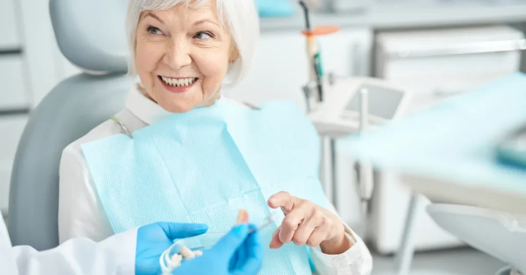 An elderly woman smiling while sitting in a dental chair, wearing a blue dental bib, and interacting with a dentist who is holding a dental model.