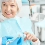 An elderly woman smiling while sitting in a dental chair, wearing a blue dental bib, and interacting with a dentist who is holding a dental model. They are discussing All-on-4 implants